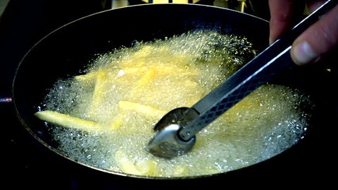 Slow motion shot of French fries being stirred by a pair of tongs in a frying pan of hot oil.