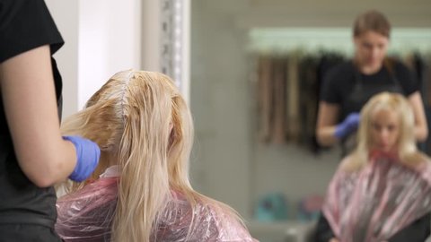 A Back View Of The Blonde Sitting In The Chair At The Hairdresser S With Dyed Hair Roots The Hairdresser Stretches Hair In Strands To Length They Are Reflected In The Mirror In Front Of Them