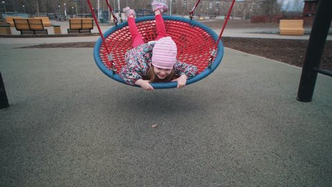 Little cute girl swinging on round swing with tight weave rope platform  seat in park on cloudy day. child laughs and waves his hand.