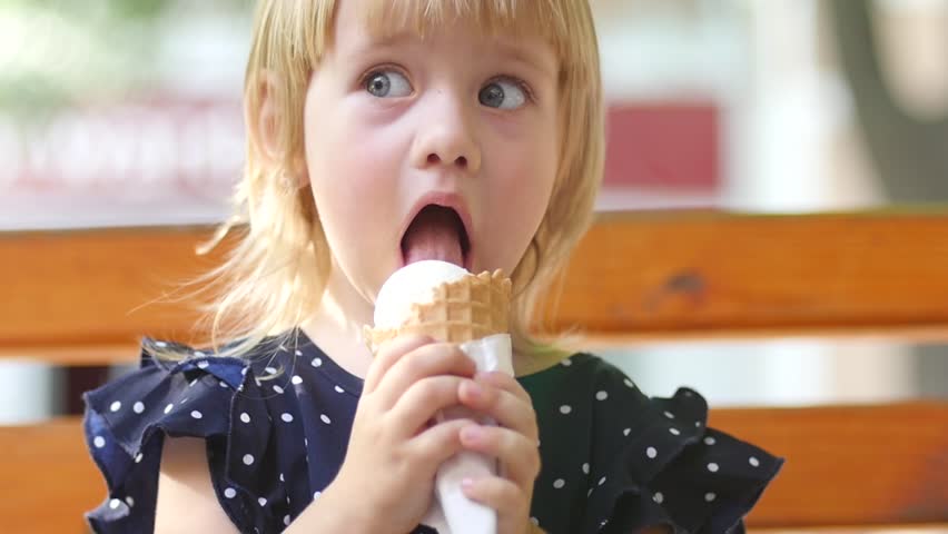 Little Blonde Girl Eats Licks An Ice Cream Cone On A Park Bench