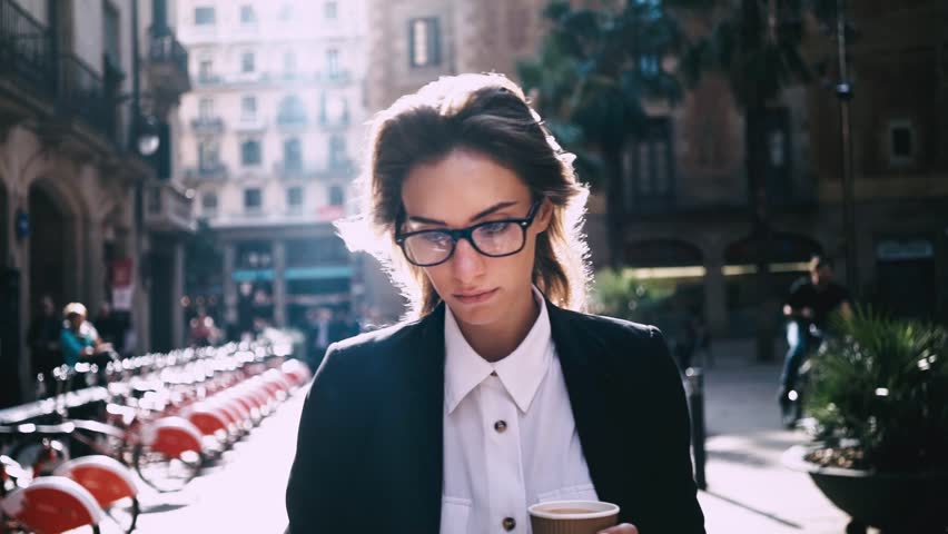 Close-up of attractive young businesswoman walking on urban street and using modern smartphone outside, sunshine, slow motion