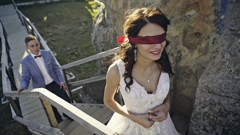 Newly married couple walking along wooden path among rocks, blindfolded bride going upstairs.